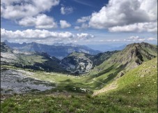 Panoramic view of a lush green alpine valley surrounded by rugged mountain peaks under a cloudy blue sky.